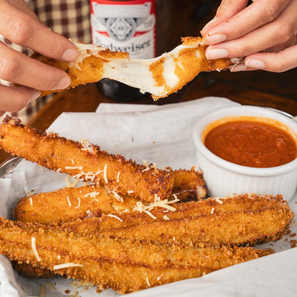 Crispy breaded cheese sticks topped with shredded cheese, one being pulled apart beside marinara dipping sauce.