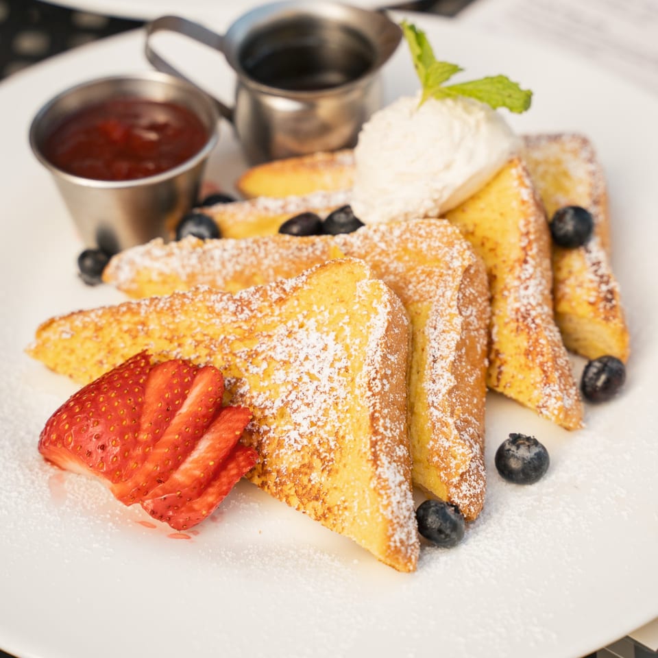 French toast dusted with powdered sugar, topped with strawberries and blueberries, served with syrup and whipped cream.