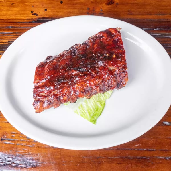 A rack of glistening BBQ ribs on a white plate with a lettuce leaf, on a wooden table.