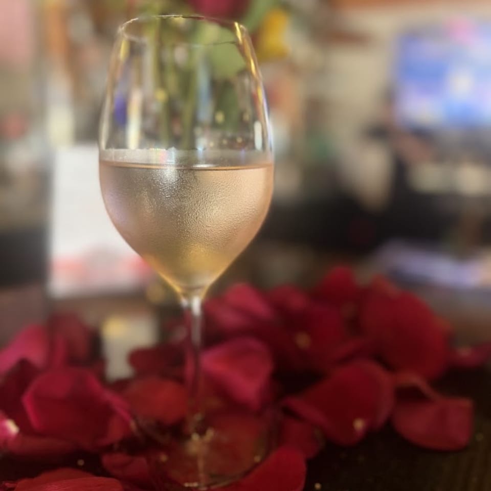 Wine glass filled with pale white wine surrounded by red rose petals on a warmly lit restaurant table.
