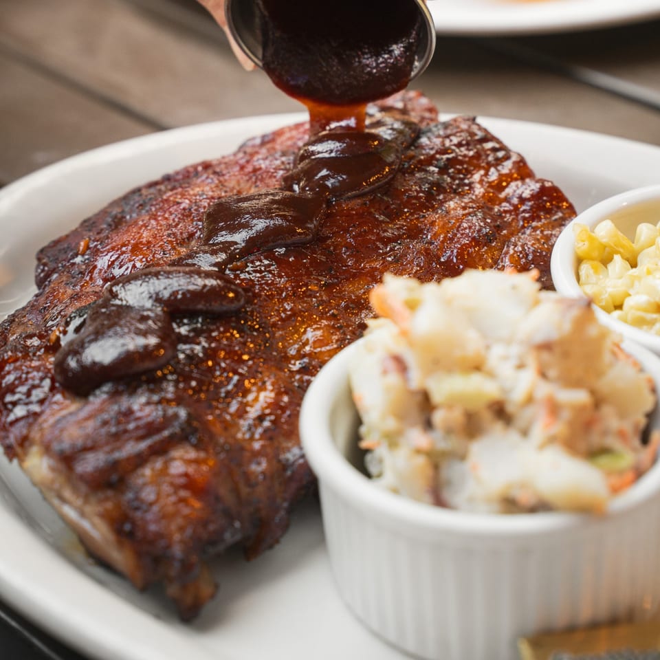 Saucy barbecue ribs topped with dark glaze, served with creamy macaroni salad and corn in white bowls.