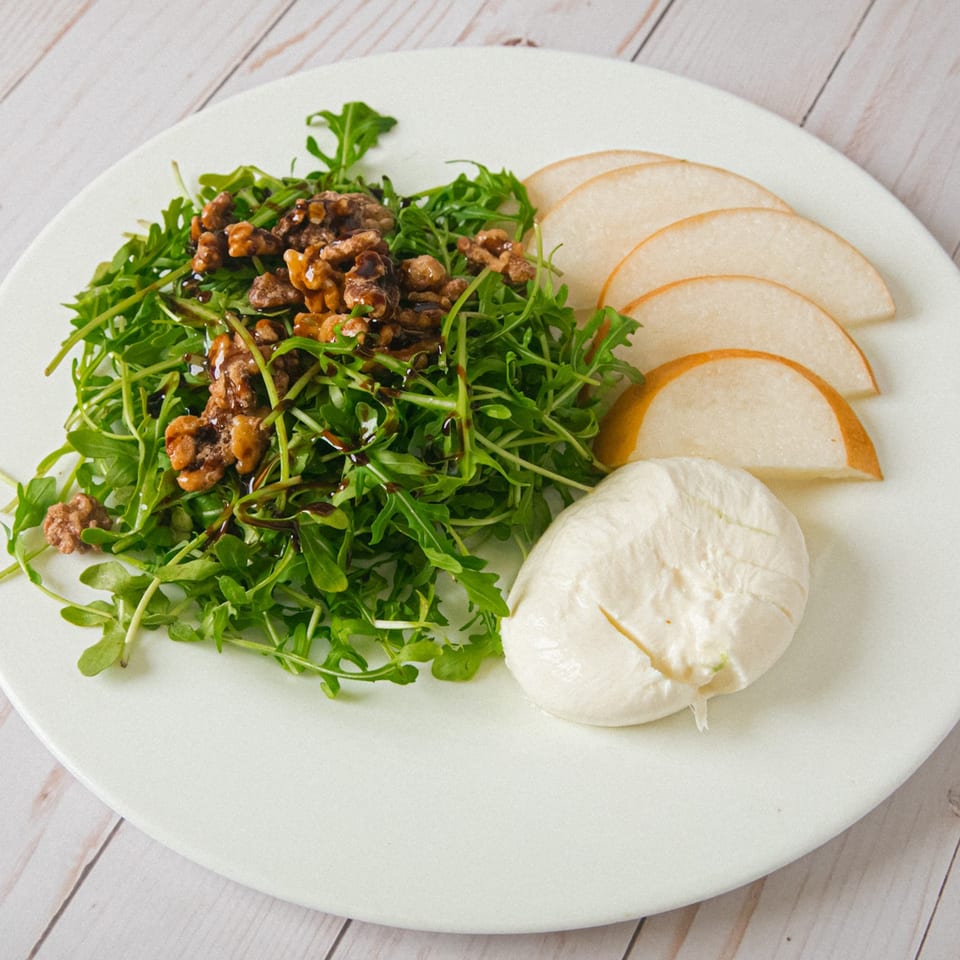 Arugula salad topped with candied walnuts and balsamic glaze, served with pear slices and creamy burrata on a white plate.