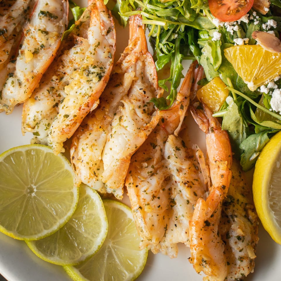 Seasoned grilled shrimp served with lime slices and a leafy citrus salad topped with crumbled cheese.
