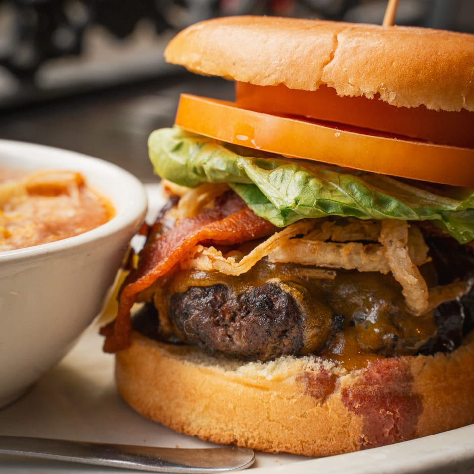 Close-up of a loaded cheeseburger with lettuce, tomato, bacon, crispy onions, and sauce, served with dipping sauce.
