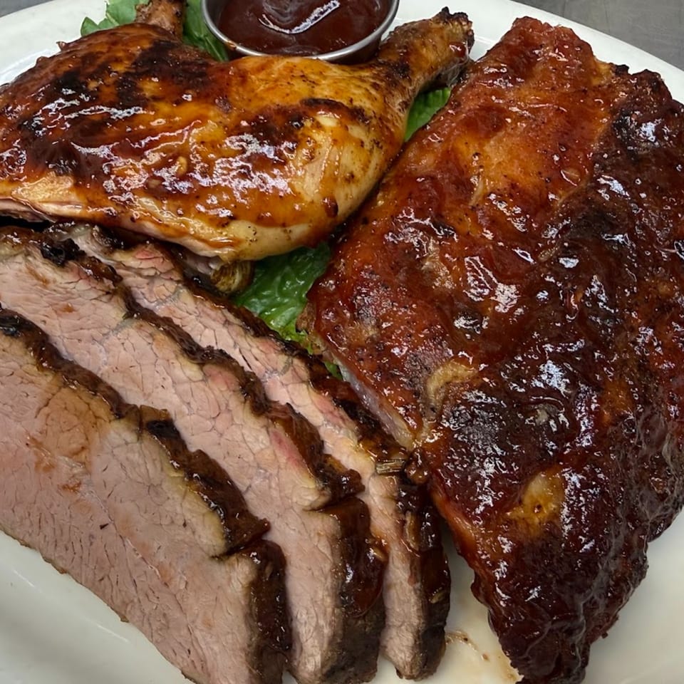 Glazed barbecue platter with sliced beef, chicken legs, ribs, leafy garnish, and dipping sauce on a white plate.