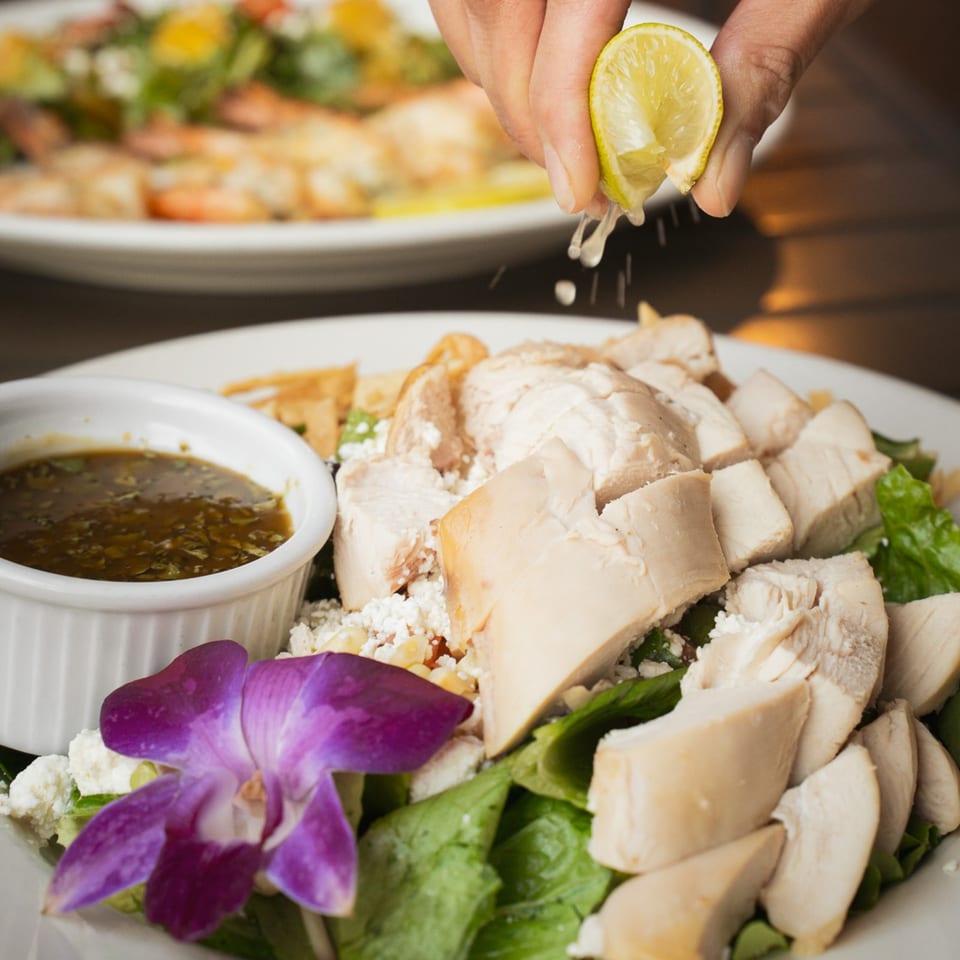 Hand squeezes lime over a platter of sliced chicken, greens, vegetables, dipping sauce, and a purple orchid garnish.