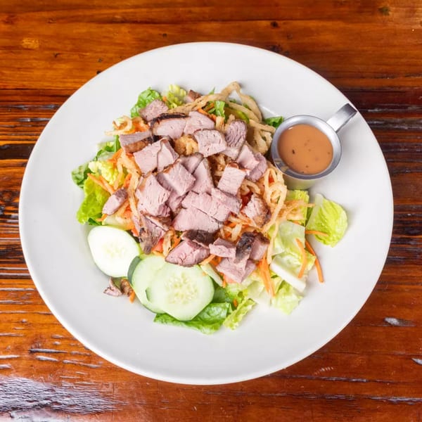 Top-down view of a steak salad with lettuce, cucumbers, carrots, crispy onions, and a side of dressing.