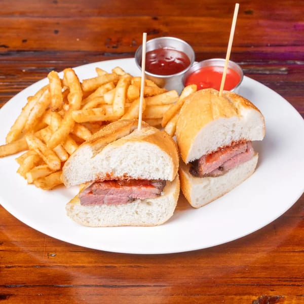 Two steak sandwiches with fries and ketchup on a white plate, on a wooden table.