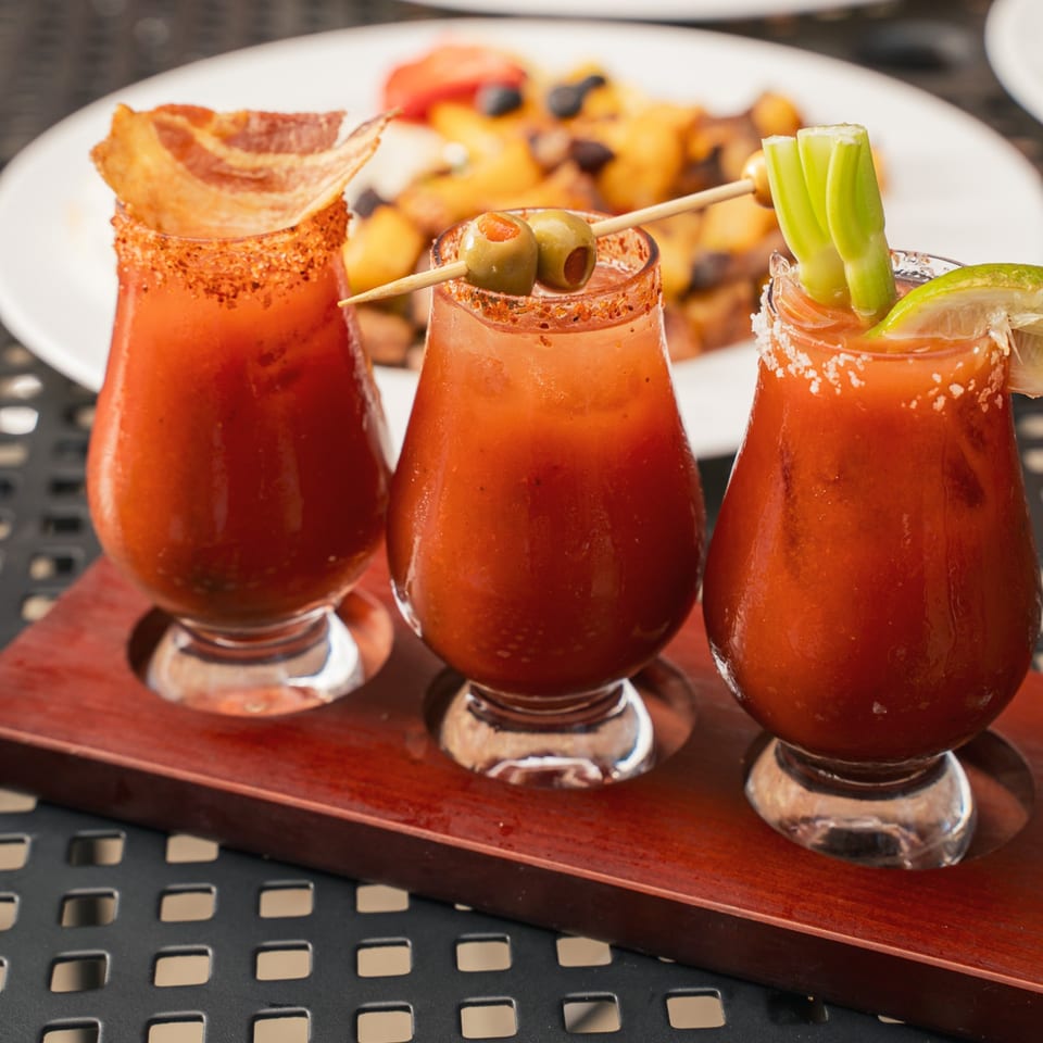 Three spicy tomato cocktails garnished with celery, lime, olives, and bacon sit on a wooden serving board beside appetizers.