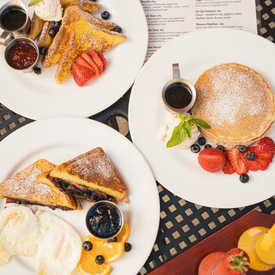 Overhead view of breakfast plates featuring French toast, pancakes, stuffed toast, fruit, whipped cream, and syrup.